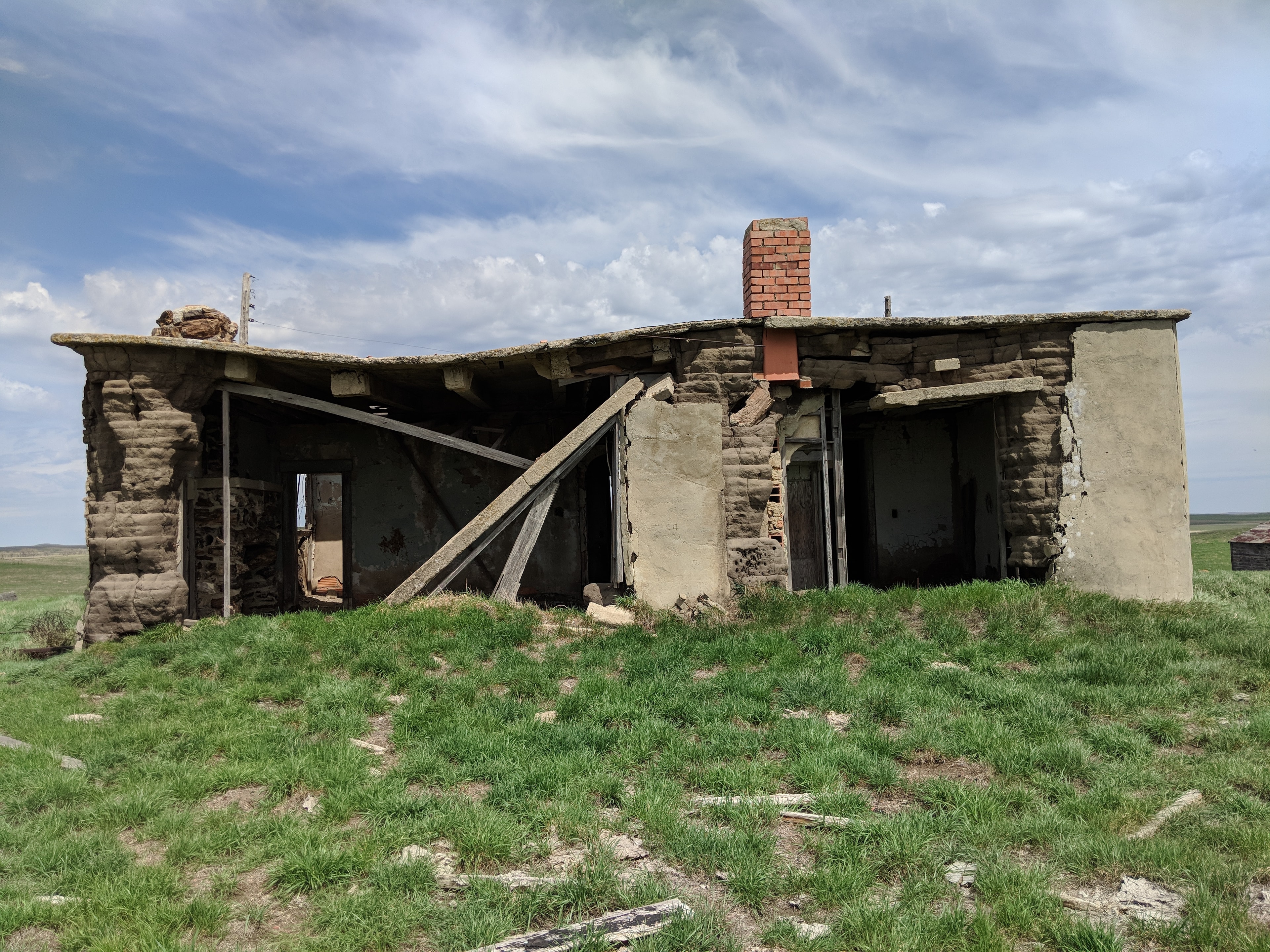 The ruins of the Scoria Lily Ranch house today — rammed earth walls partially collapsed, brick chimney still standing, on the open North Dakota prairie.
