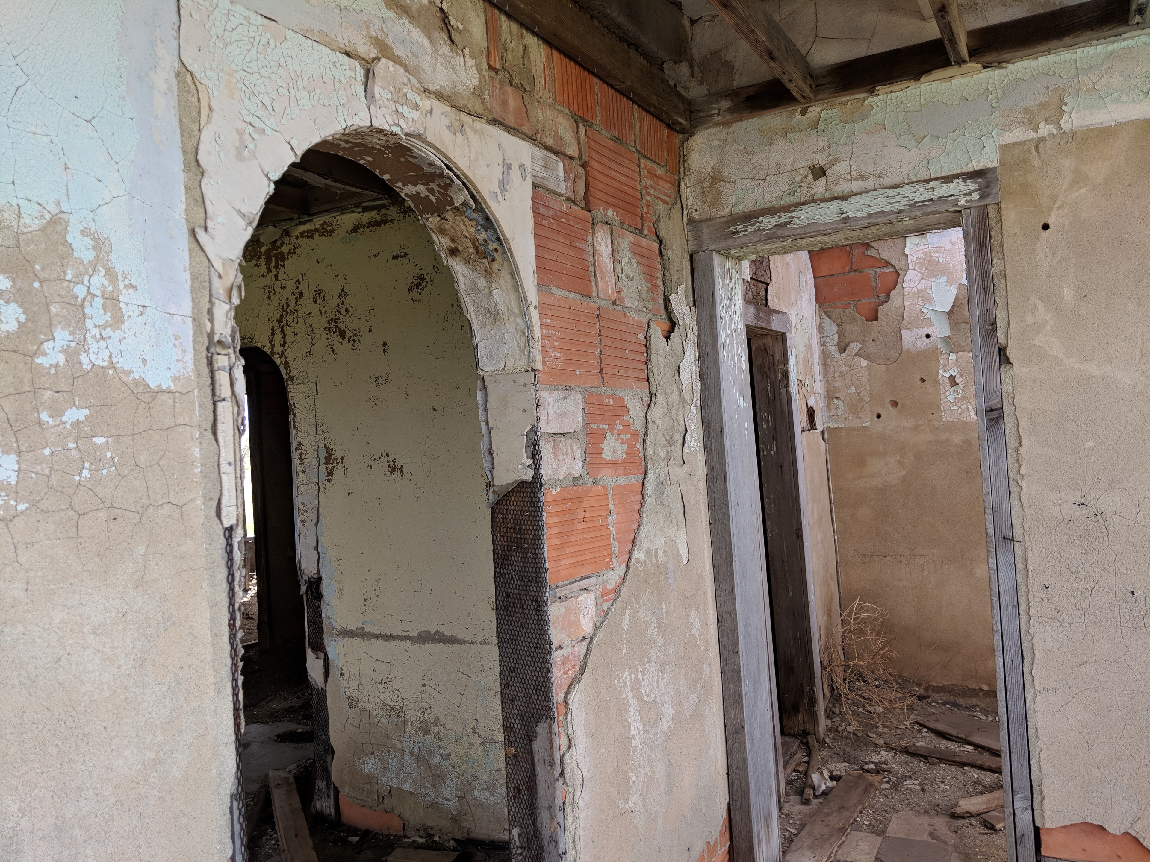 Interior of the ruined Bliss rammed-earth house — a graceful arched doorway with peeling plaster and exposed brick, debris on the floor, open sky visible through the openings.