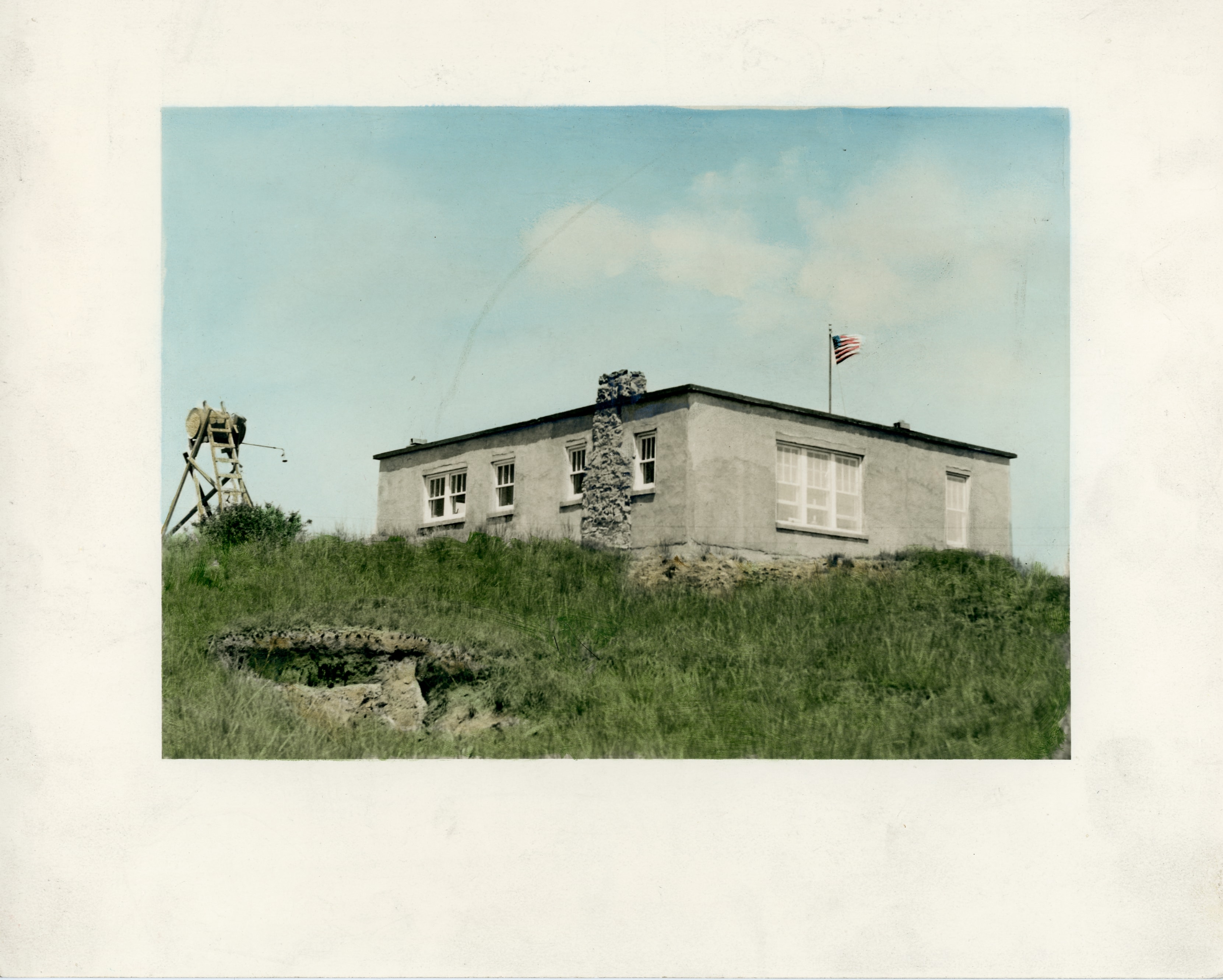 The Scoria Lily ranch house on a grassy hilltop, a low stucco building with a stone chimney and American flag, with a windmill tower to the left, 1937.